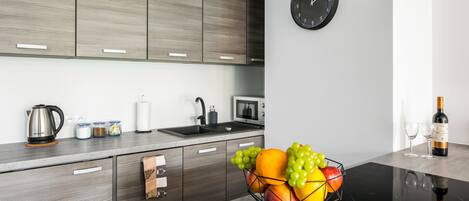 A modern kitchen with wooden cabinets, a black clock, a fruit bowl, a kettle, a microwave, and wine glasses.