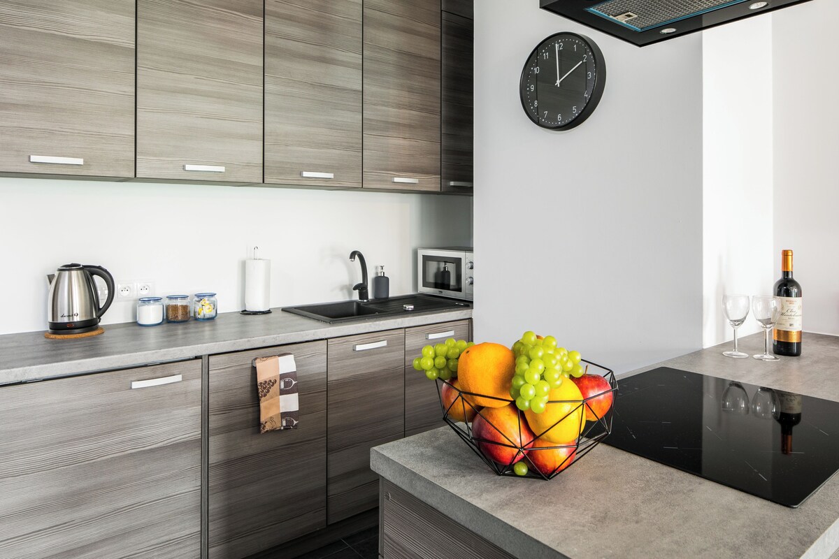 A modern kitchen with wooden cabinets, a black induction cooktop, a fruit bowl, a kettle, and a wine bottle.