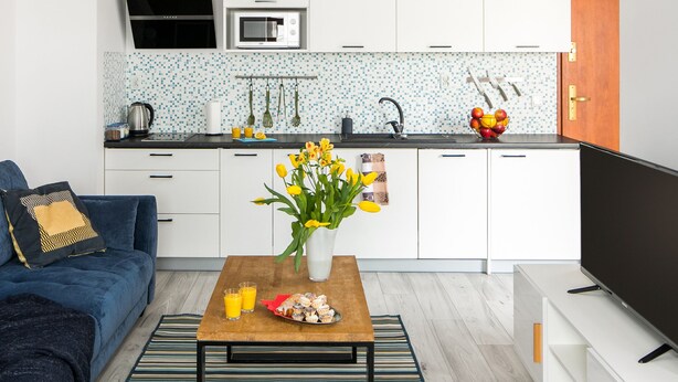 A modern kitchen with white cabinets, a black countertop, and a tiled backsplash. There is a blue sofa with a decorative pillow, a wooden coffee table with a vase of flowers, and a flat-screen TV. - Green Garden Residence (Kraków)