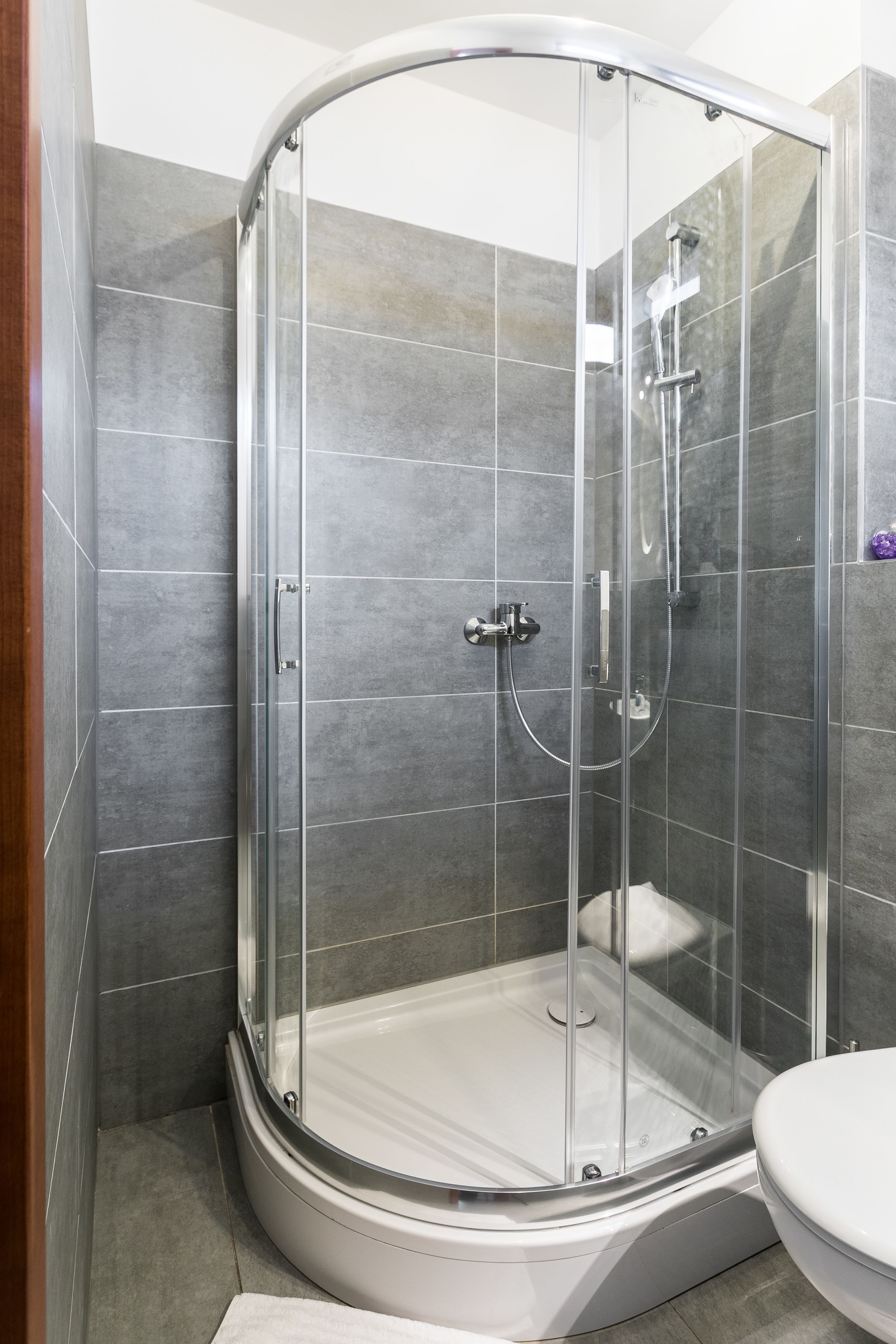 A glass-enclosed shower stall with a white bathtub, grey tiled walls, and a white toilet.
