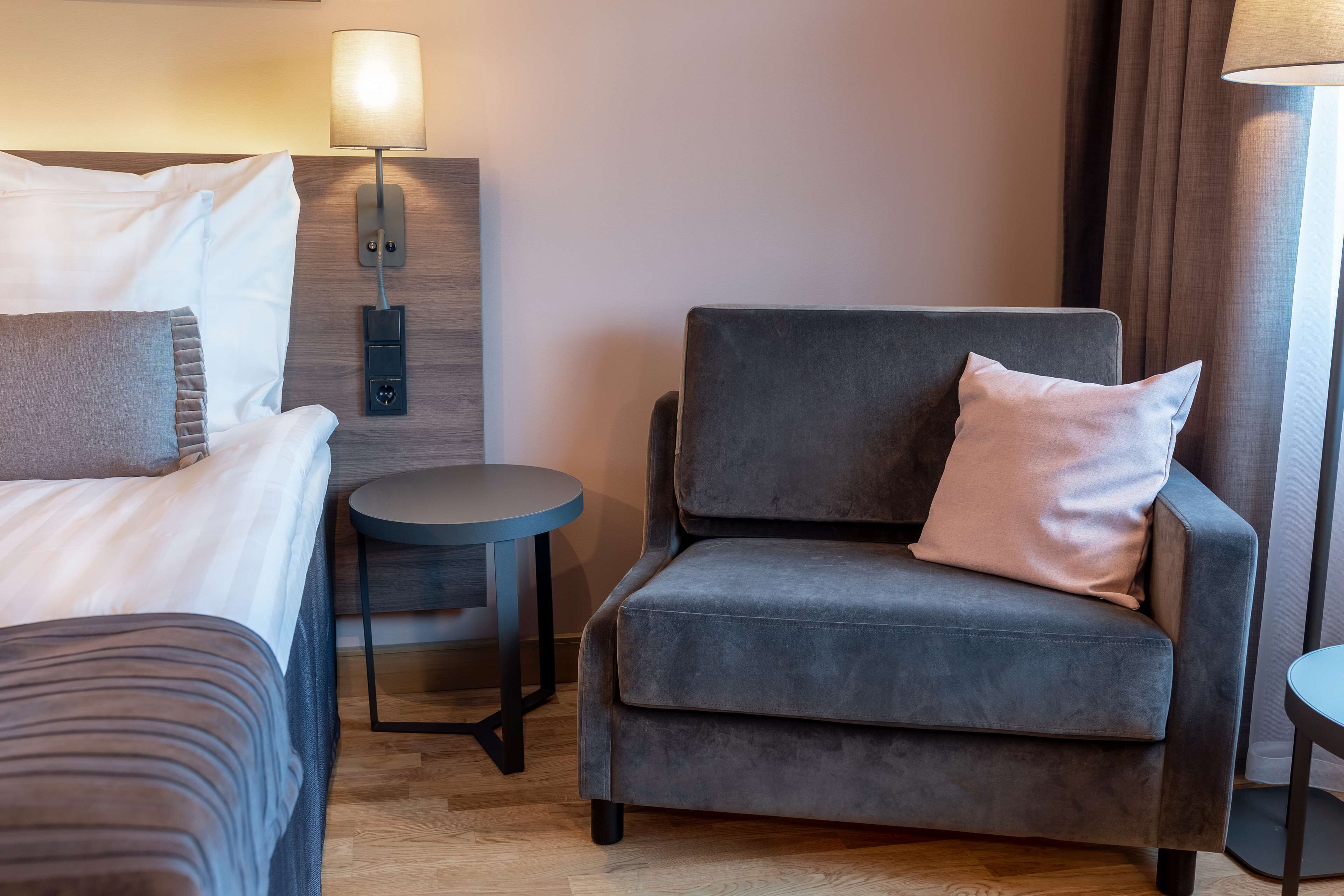 Une chambre d’hôtel avec un lit, un fauteuil gris, une petite table ronde et une lampe fixée au mur.