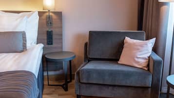 Hotellihuone, jossa on sänky, harmaa nojatuoli, pieni pyöreä pöytä ja seinään kiinnitetty lamppu.