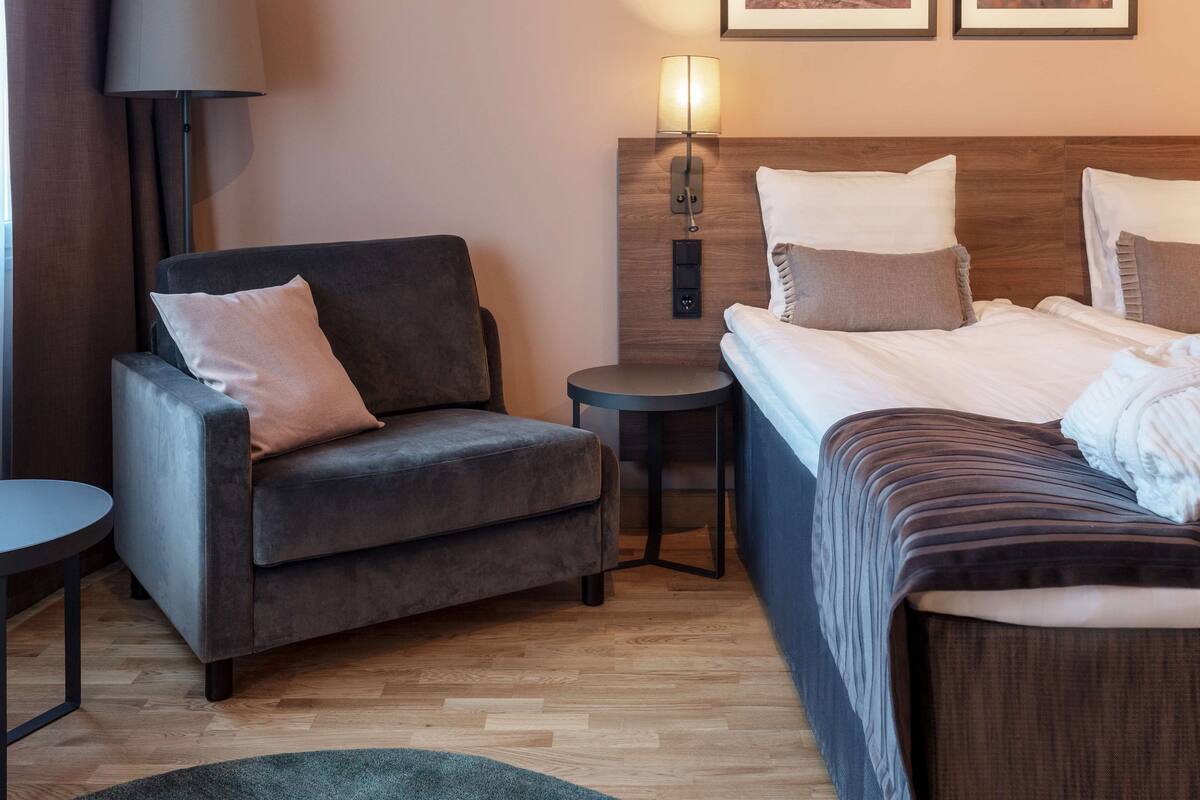 A hotel room with a sofa, a small round table, a bed with a wooden headboard, and a wall-mounted lamp.