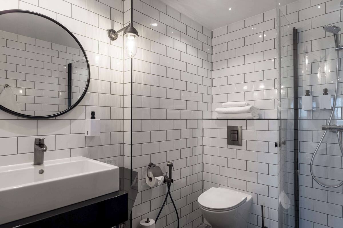 A modern bathroom with a white tiled wall, a round mirror, a sink, a toilet, and a glass-enclosed shower area.