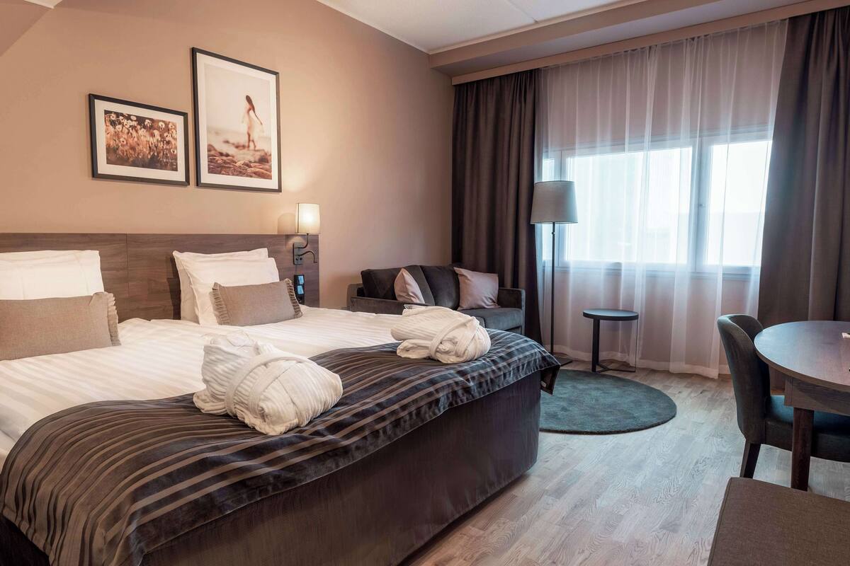 A hotel room with a large bed, a desk, a chair, a small table, and a window with curtains.