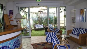 Zona de recepción con suelo de baldosas, un mostrador de recepción, muebles de mimbre y vistas a un jardín exterior.