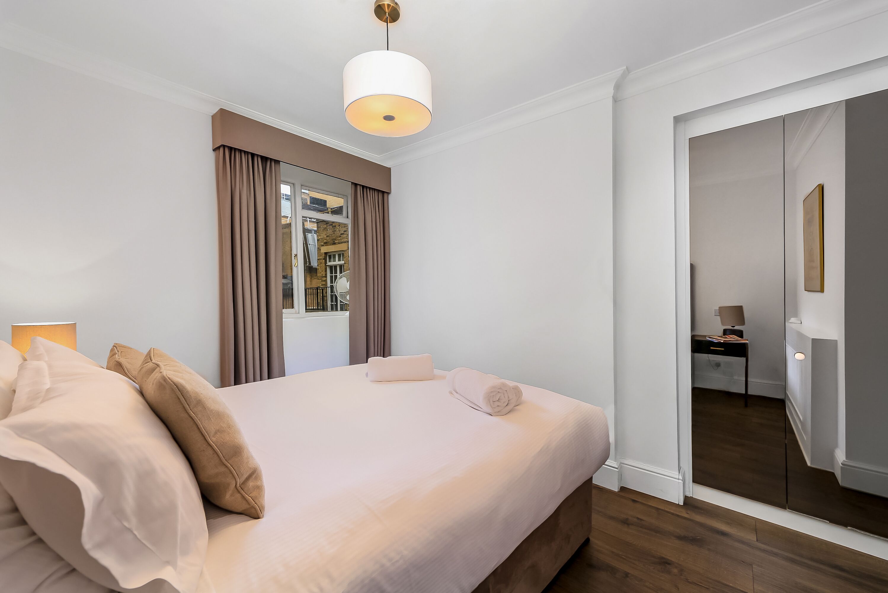 Una cama bien tendida con ropa de cama y almohadas color beige, una lámpara de noche y una ventana con cortinas.