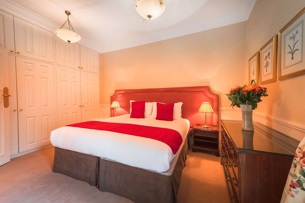 A hotel room with a large bed, two bedside tables, a nightstand, a dresser, a vase with flowers, and framed pictures on the wall.