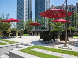 An outdoor seating area with red umbrellas, tables, and chairs, surrounded by greenery and modern buildings in the background.