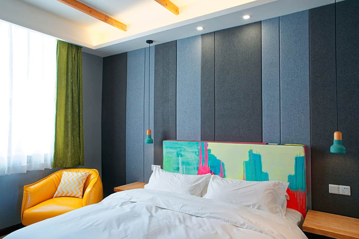 A hotel room with a bed, a chair, a colorful headboard, and a window with curtains.