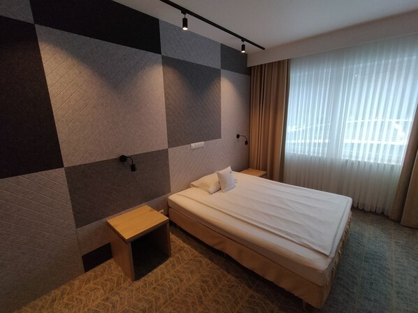 Una habitación de hotel con una cama grande, una mesita de noche, una lámpara de pared y una ventana con cortinas.