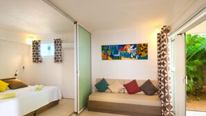 Habitación de hotel con cama, sofá, un cuadro abstracto colorido y puerta corrediza de vidrio que da a un jardín.