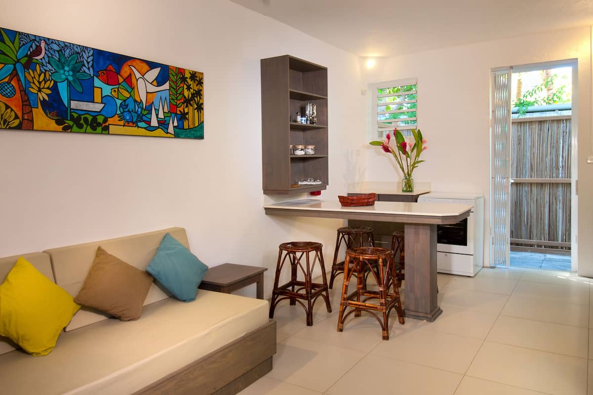 A living room with a sofa, a coffee table, and a kitchen area with a bar stool and a vase of flowers.