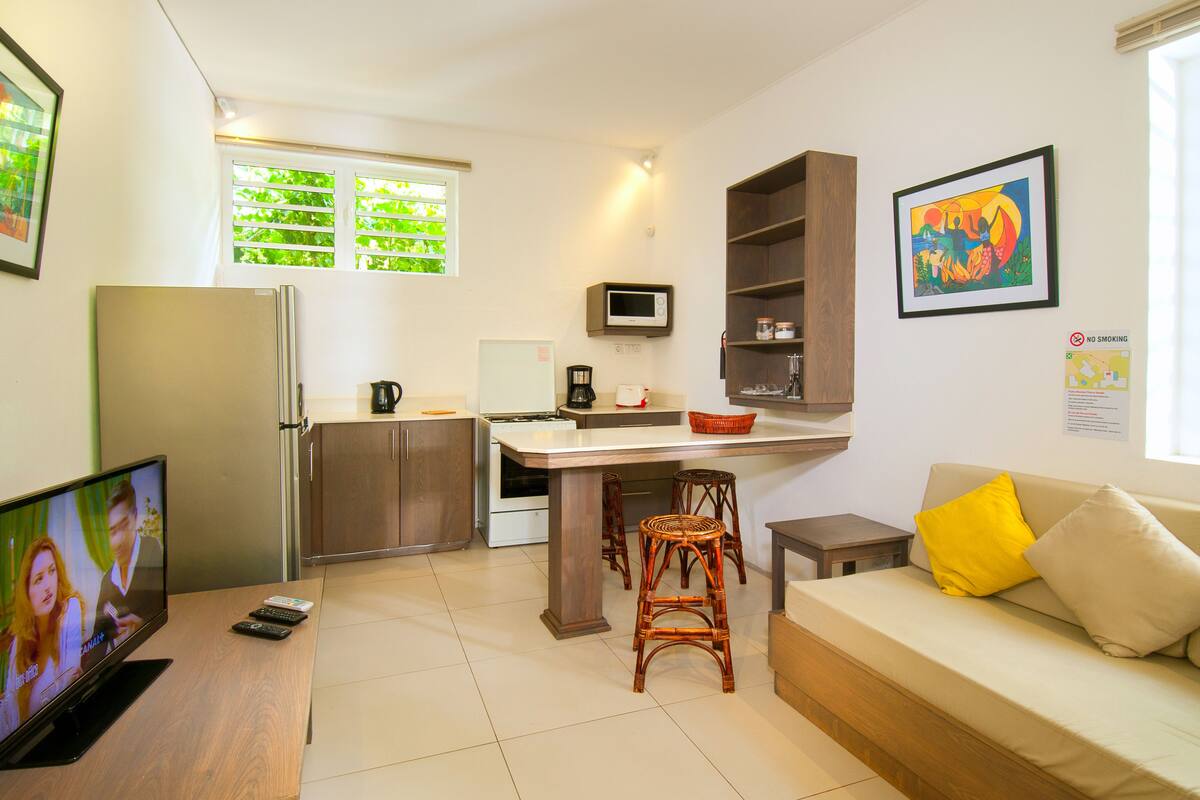A compact kitchen with a refrigerator, microwave, and coffee maker. There is a dining area with a table and chairs, and a sofa with cushions.