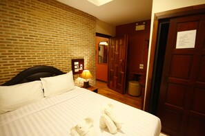 A hotel room with a brick wall, a bed with white linens, a nightstand with a lamp, a wooden wardrobe, and a mirror.