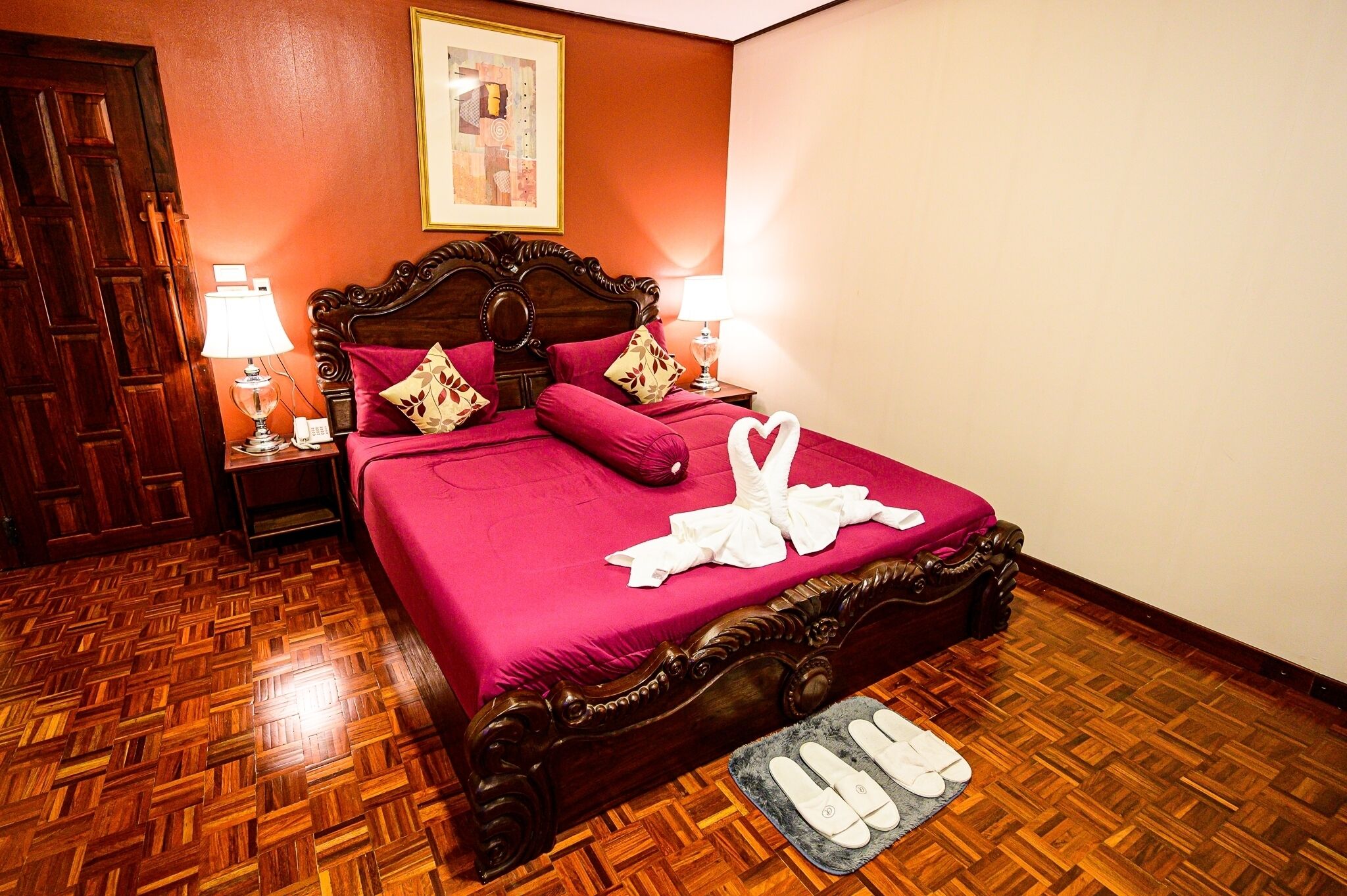 A hotel room with a wooden parquet floor, a large bed with a wooden headboard, two bedside lamps, and a framed artwork on the wall.