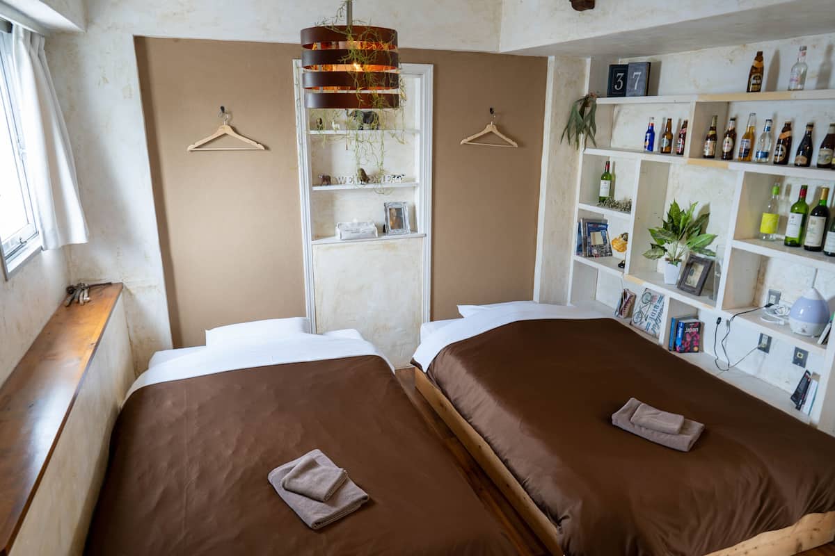 A hotel room with two beds, a bookshelf, and a window with a curtain.