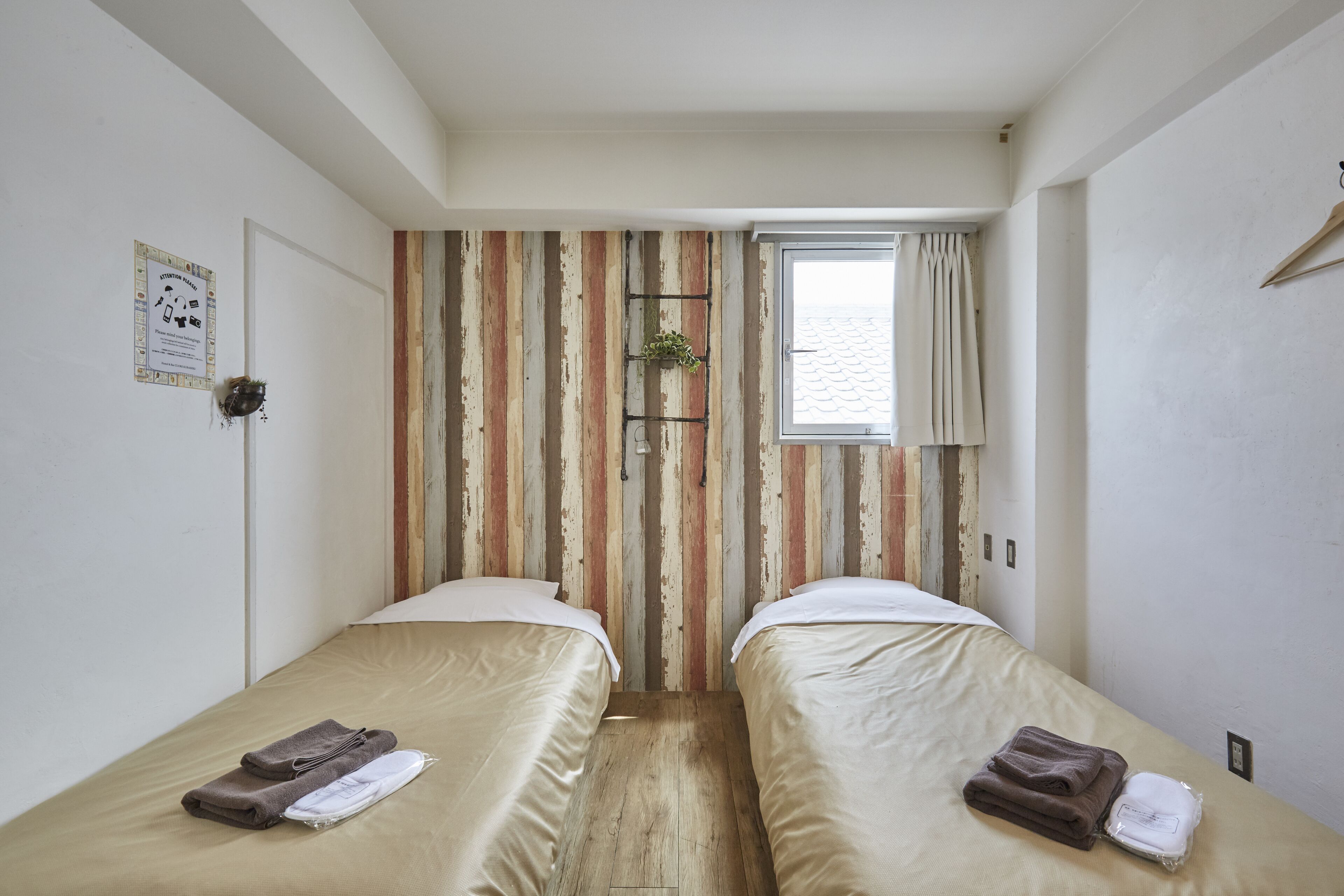 A small, clean room with two beds, a window with curtains, and a wooden wall with a striped pattern.
