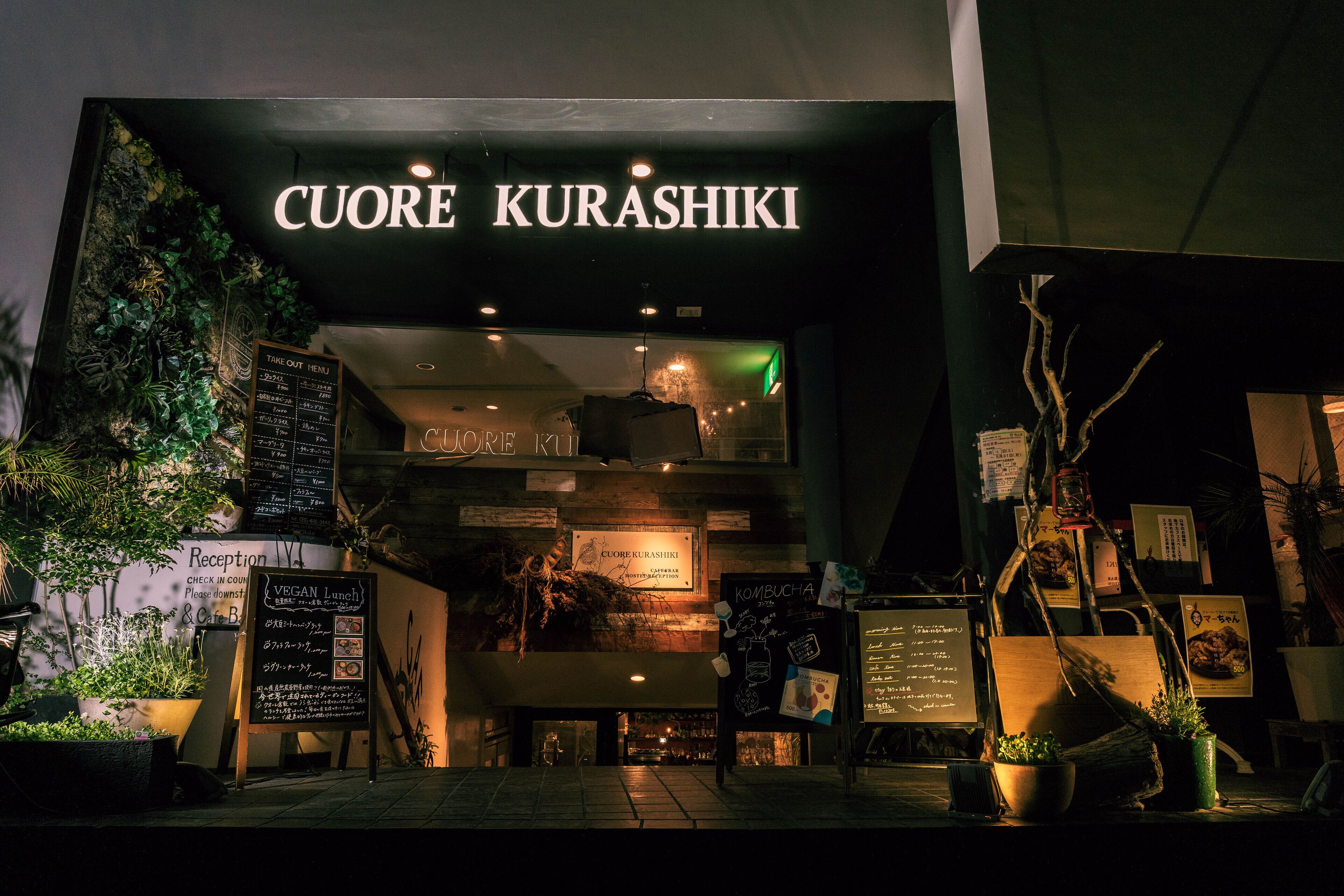 Exterior do restaurante chamado "CUORE KURASHIKI", com letreiro, cardápio e alguns vasos de plantas.