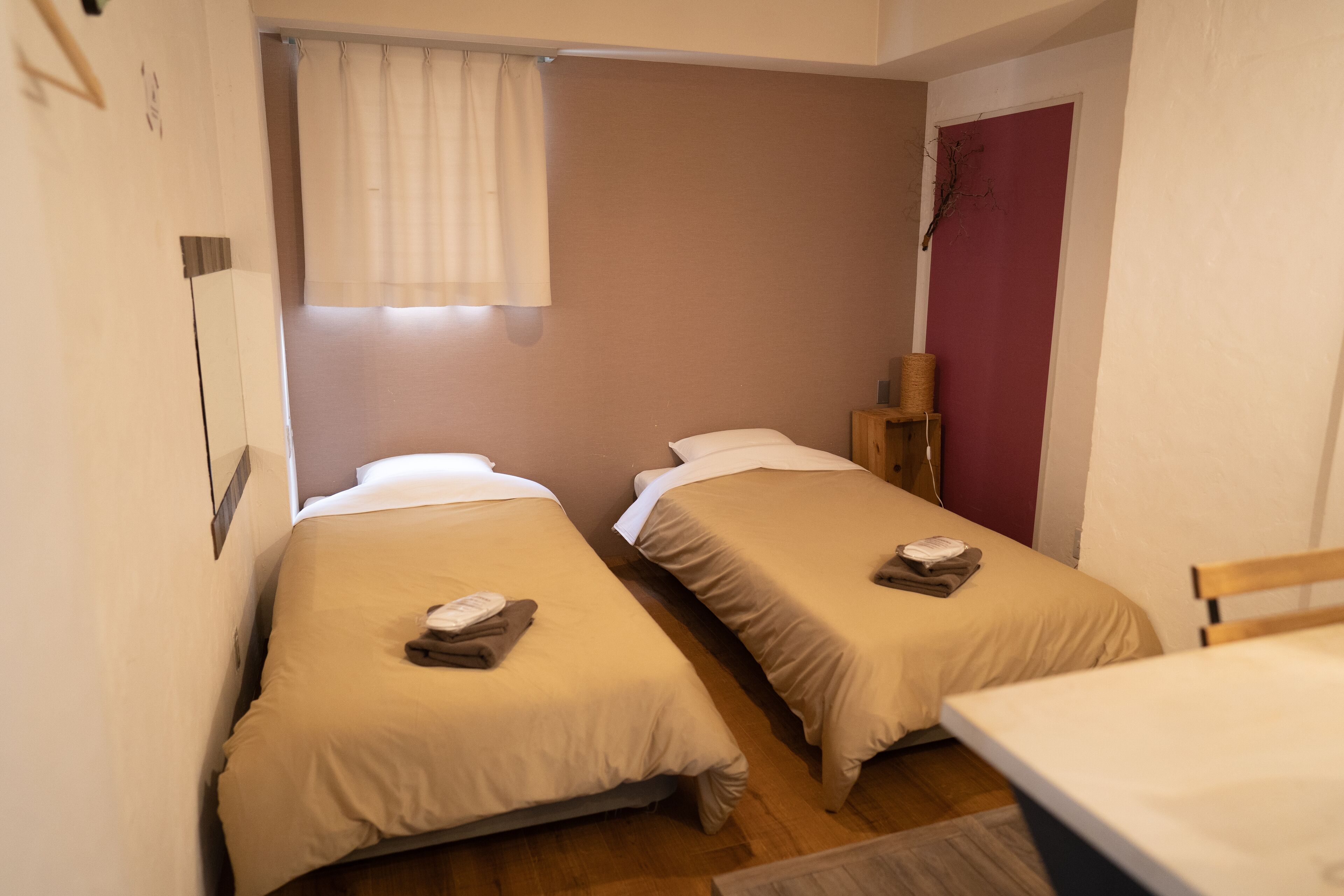 A small, compact room with two single beds, a wooden chair, a small table, and a window with curtains.