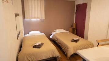 A small, compact room with two single beds, a wooden chair, a small table, and a window with curtains.