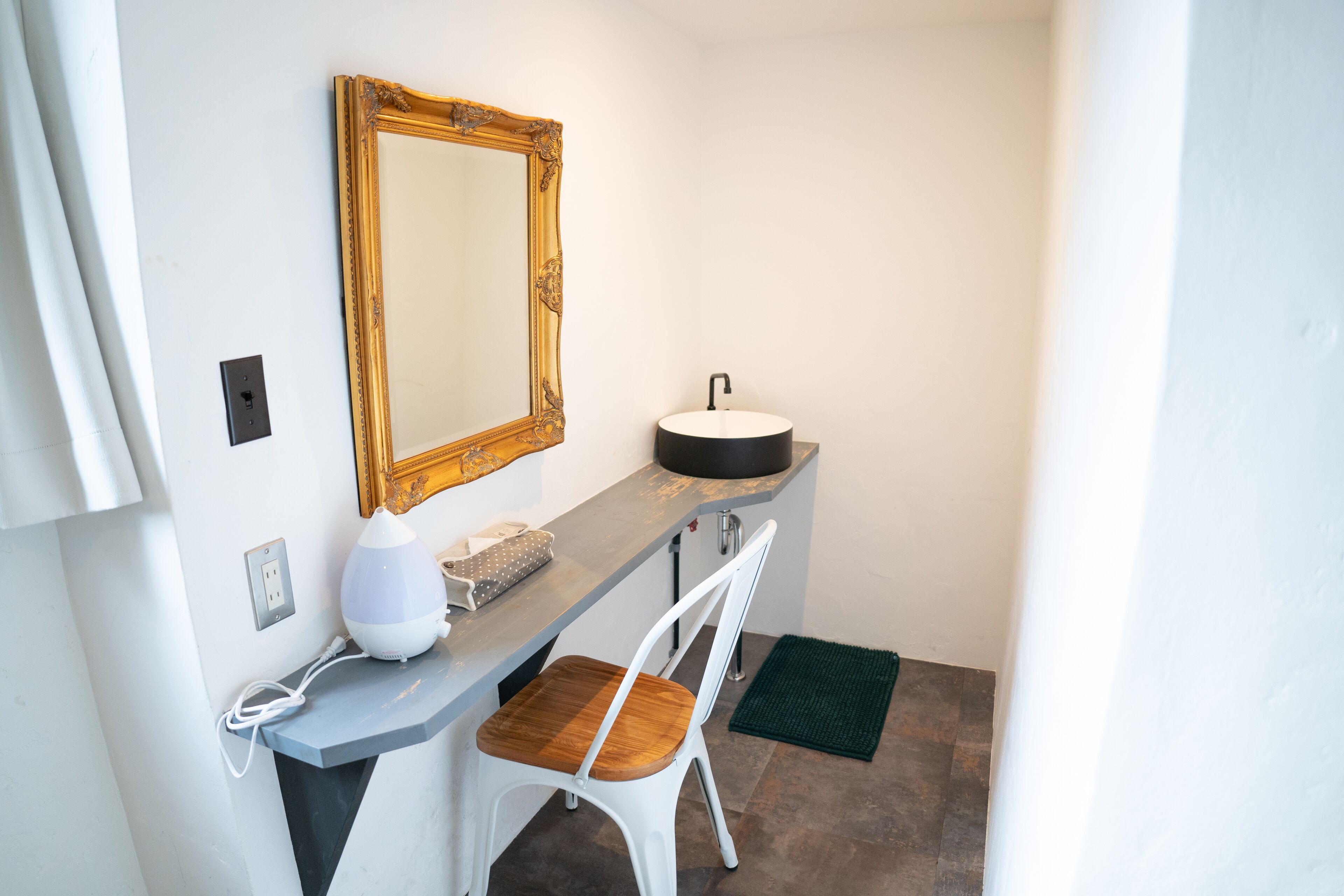 A compact bathroom with a white sink, a wooden chair, a mirror with an ornate frame, and a green mat on the floor.