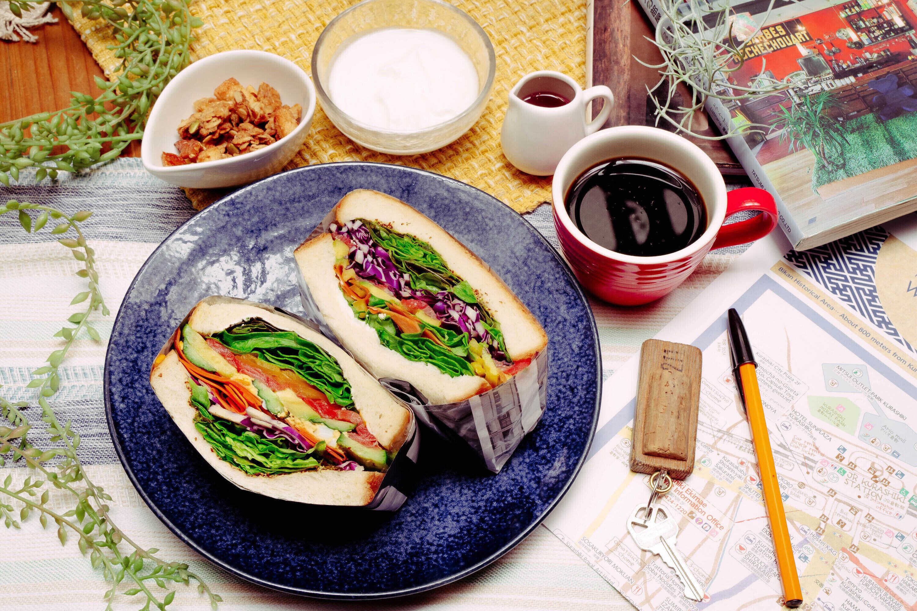 A plate with two sandwiches, a cup of coffee, a bowl of yogurt, and a book on a table.