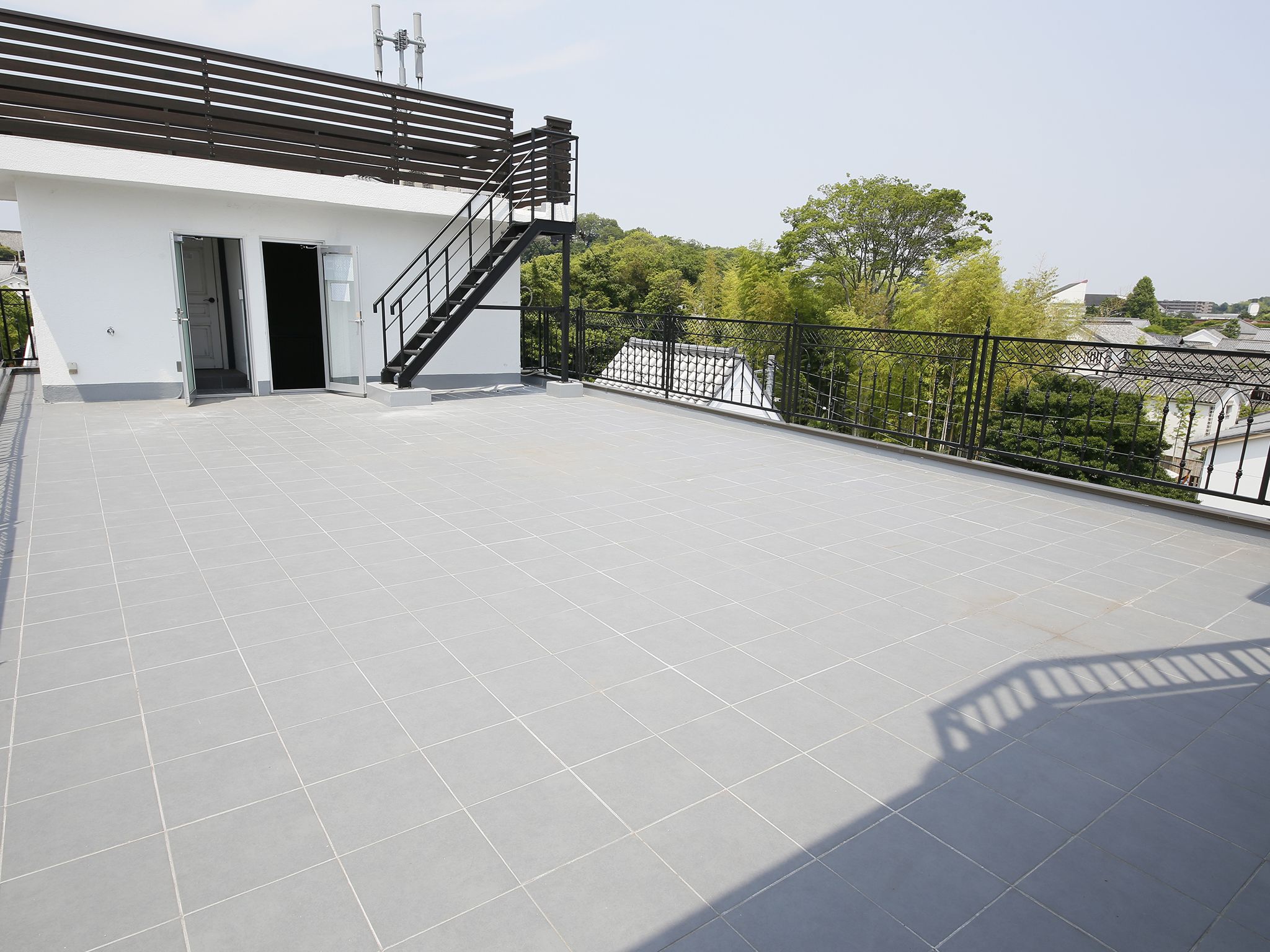 A rooftop terrace with a modern design, featuring a staircase leading to an entrance, surrounded by a metal railing and a view of greenery and buildings in the background.