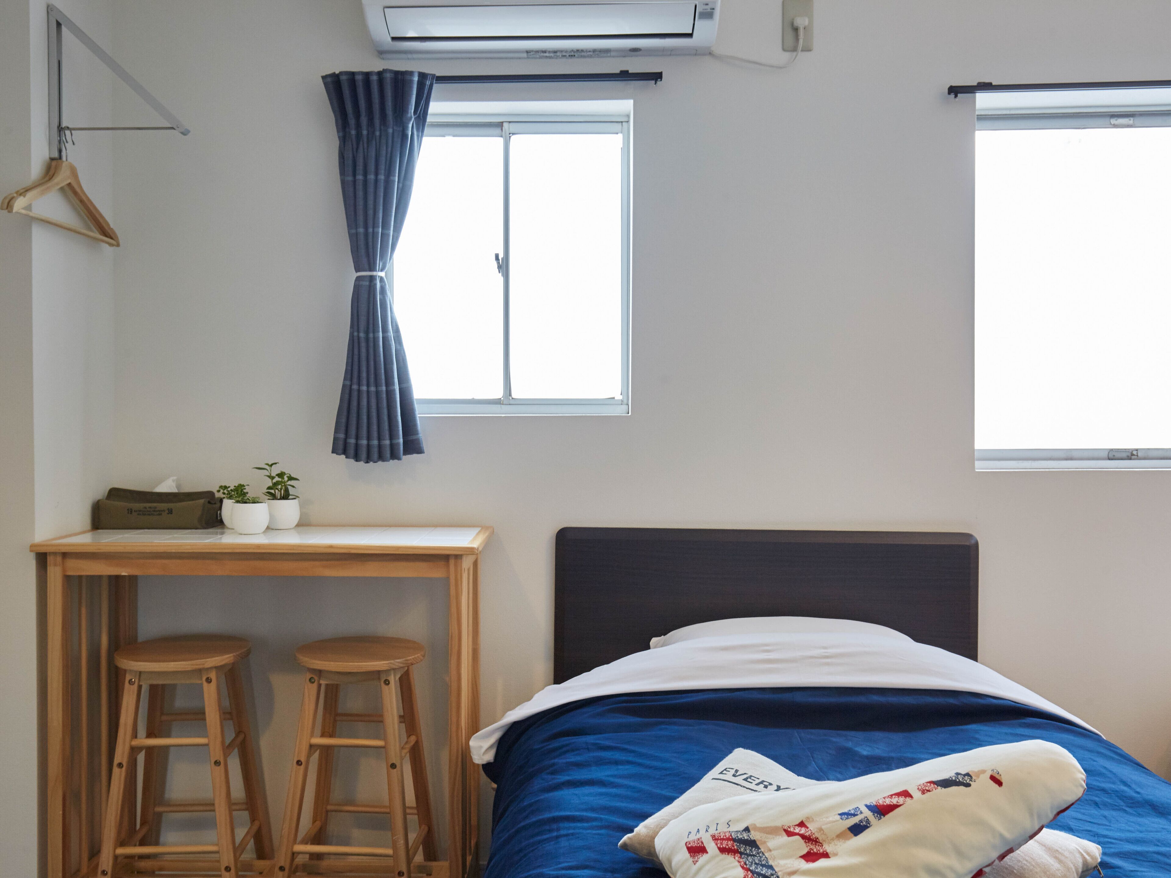 A bedroom with a bed, a desk with two stools, a window with curtains, and an air conditioner.