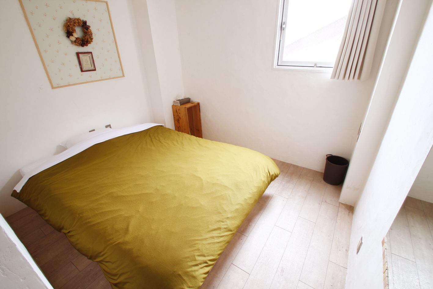 Een slaapkamer met een bed, een klein houten krukje, een prullenbak en een lijst met een afbeelding aan de muur.