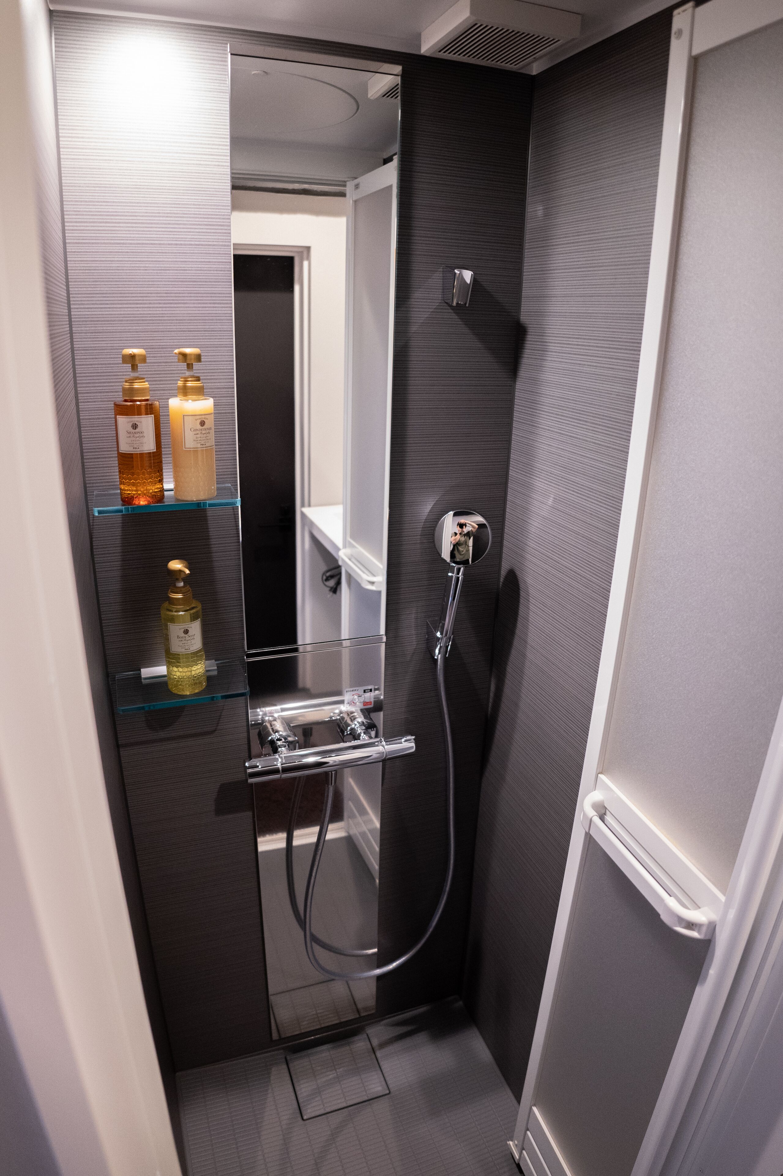 A modern bathroom with a glass shelf holding toiletries, a shower with a handheld showerhead, and a mirror.