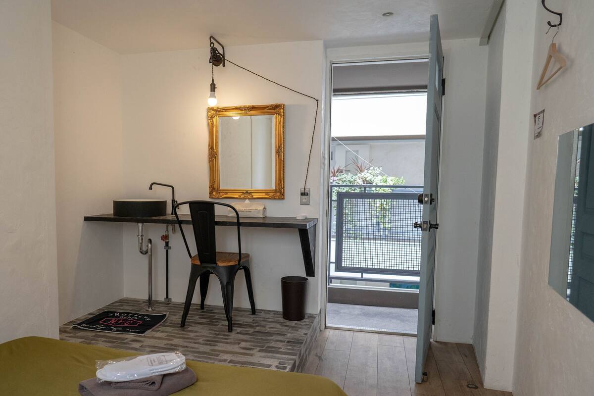 A bedroom with a bed, a desk, a chair, a mirror, and a view of the outside.