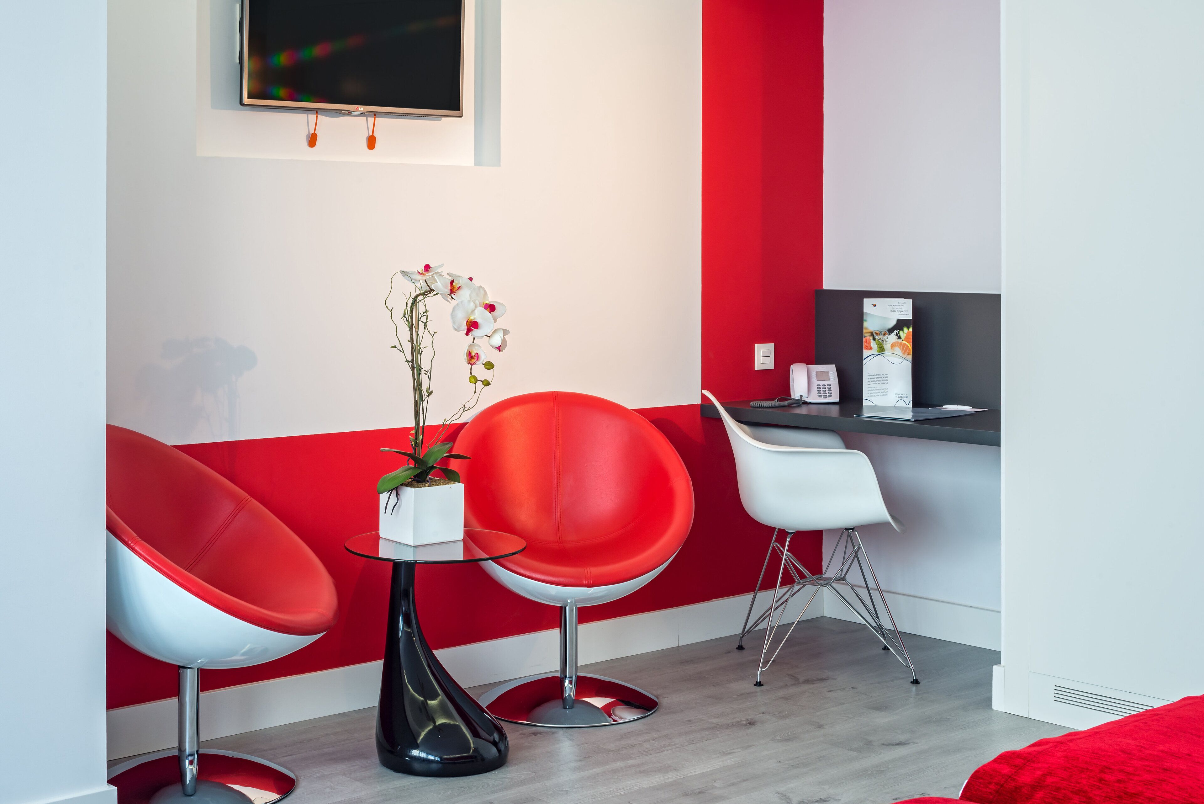 A modern room with red and white walls, two red chairs, a small table with a plant, and a desk with a computer and a phone.