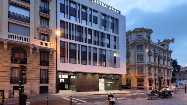 Un hotel con fachada moderna, un edificio clásico a la derecha y una calle en primer plano.