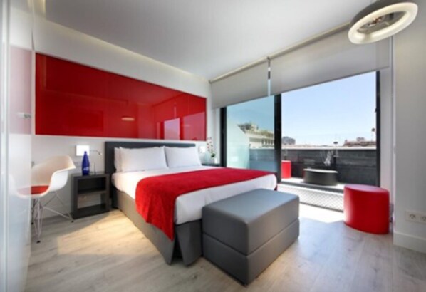 Un dormitorio moderno con una cama grande, una pared de acento roja, un reposapiés gris, una silla blanca y un balcón con un taburete rojo.