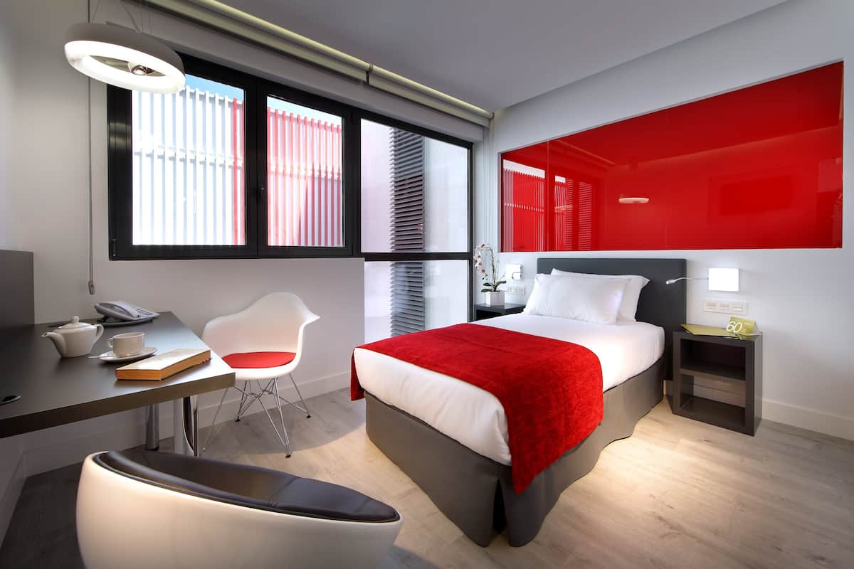 A modern hotel room with a bed, desk, chair, and a red accent wall.