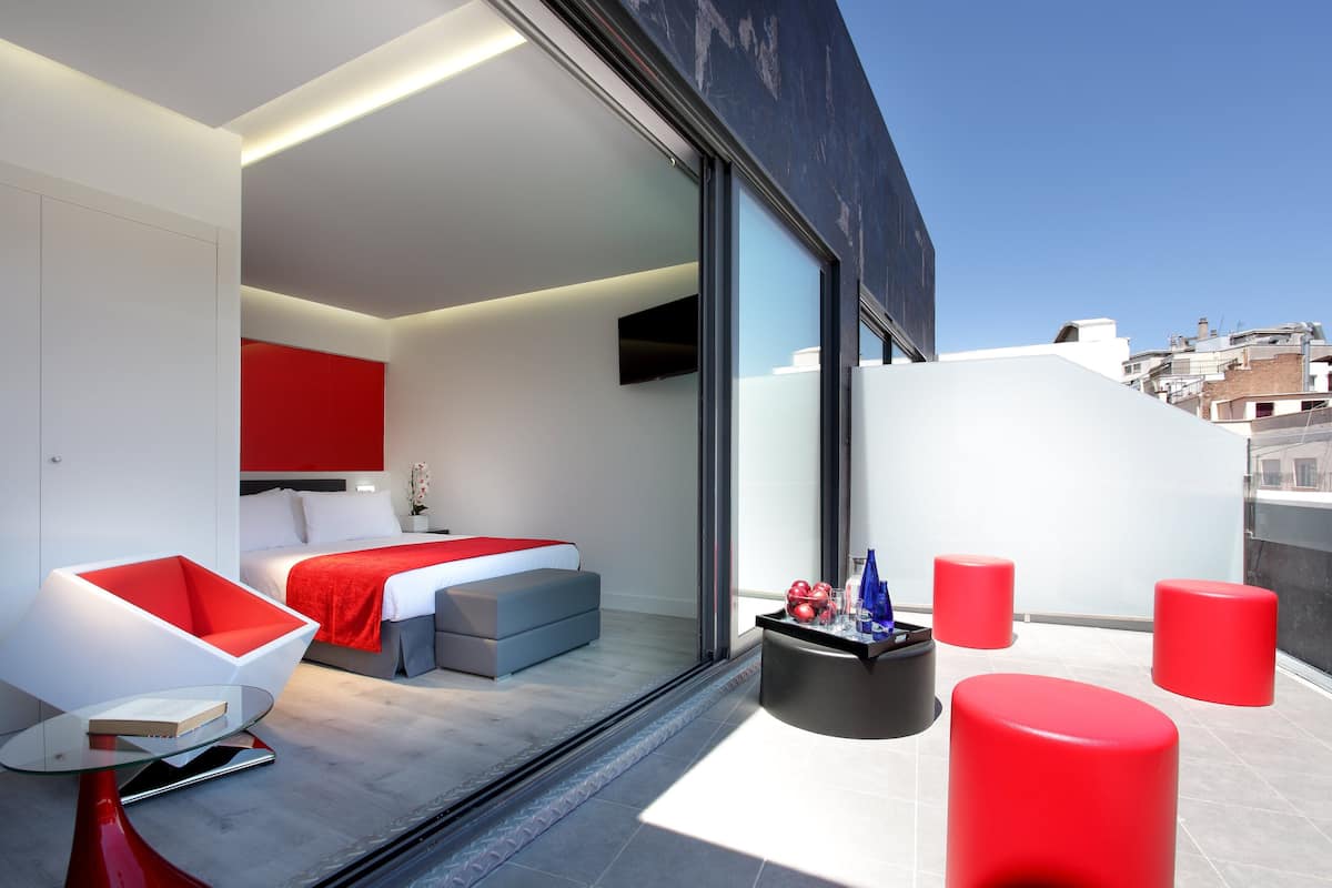 A modern hotel room with a large bed, a red accent wall, a glass partition, and a balcony with red stools.