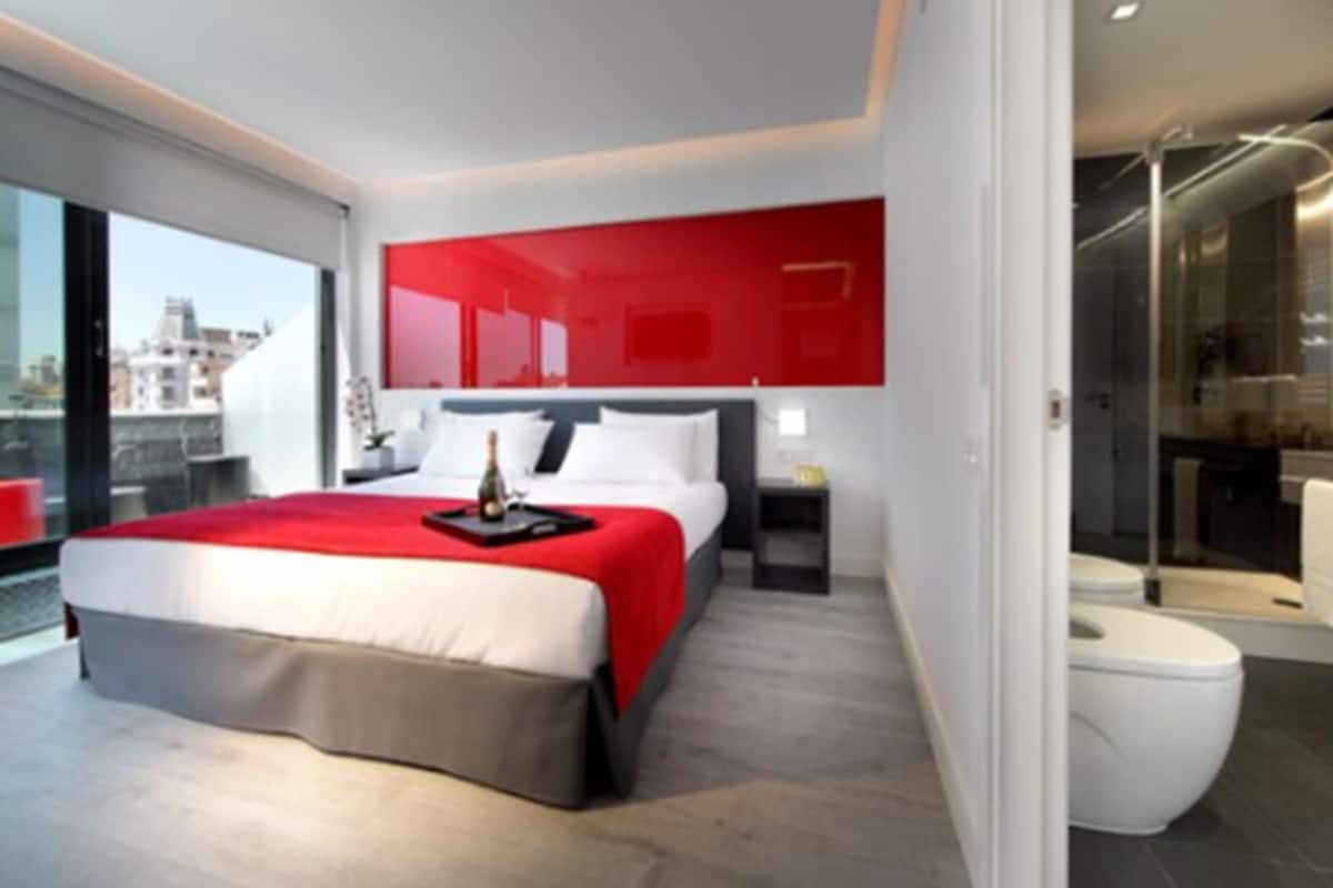 A modern hotel room with a large bed, a red accent wall, and a city view.