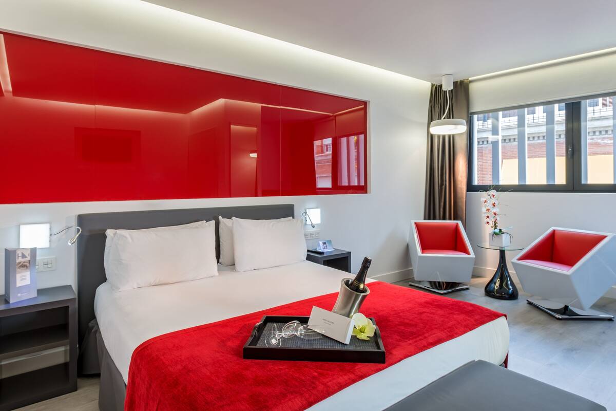 A modern hotel room with a large bed, red accents, a minibar, and a view of the city.