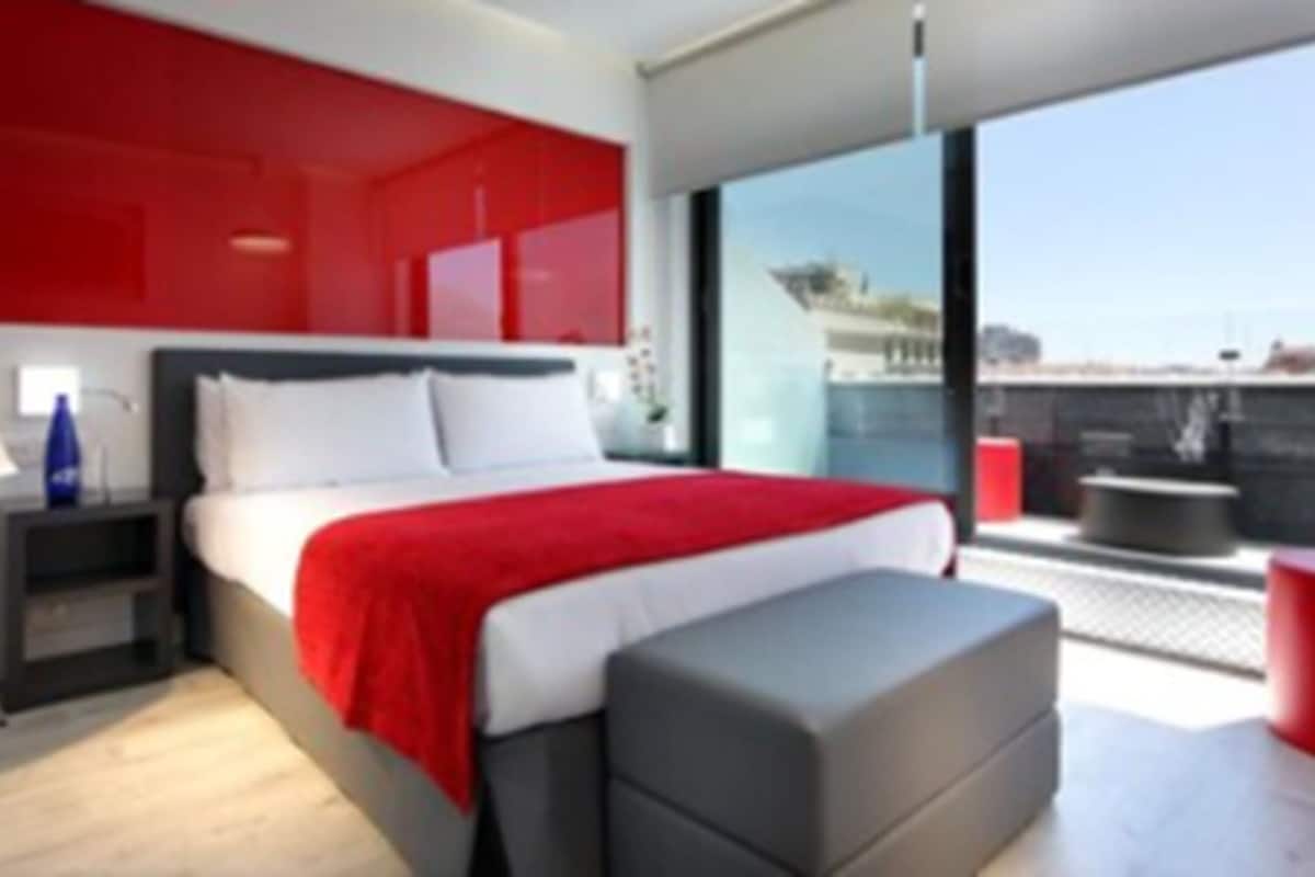 A modern bedroom with a large bed, a red accent wall, a balcony with a red stool, and a view of the city.