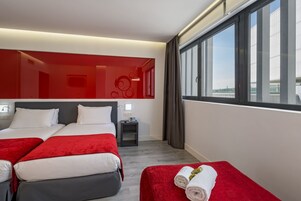 Habitación de hotel con dos camas, una pared de acento roja, una mesita de noche con lámpara y un ventanal con vistas.