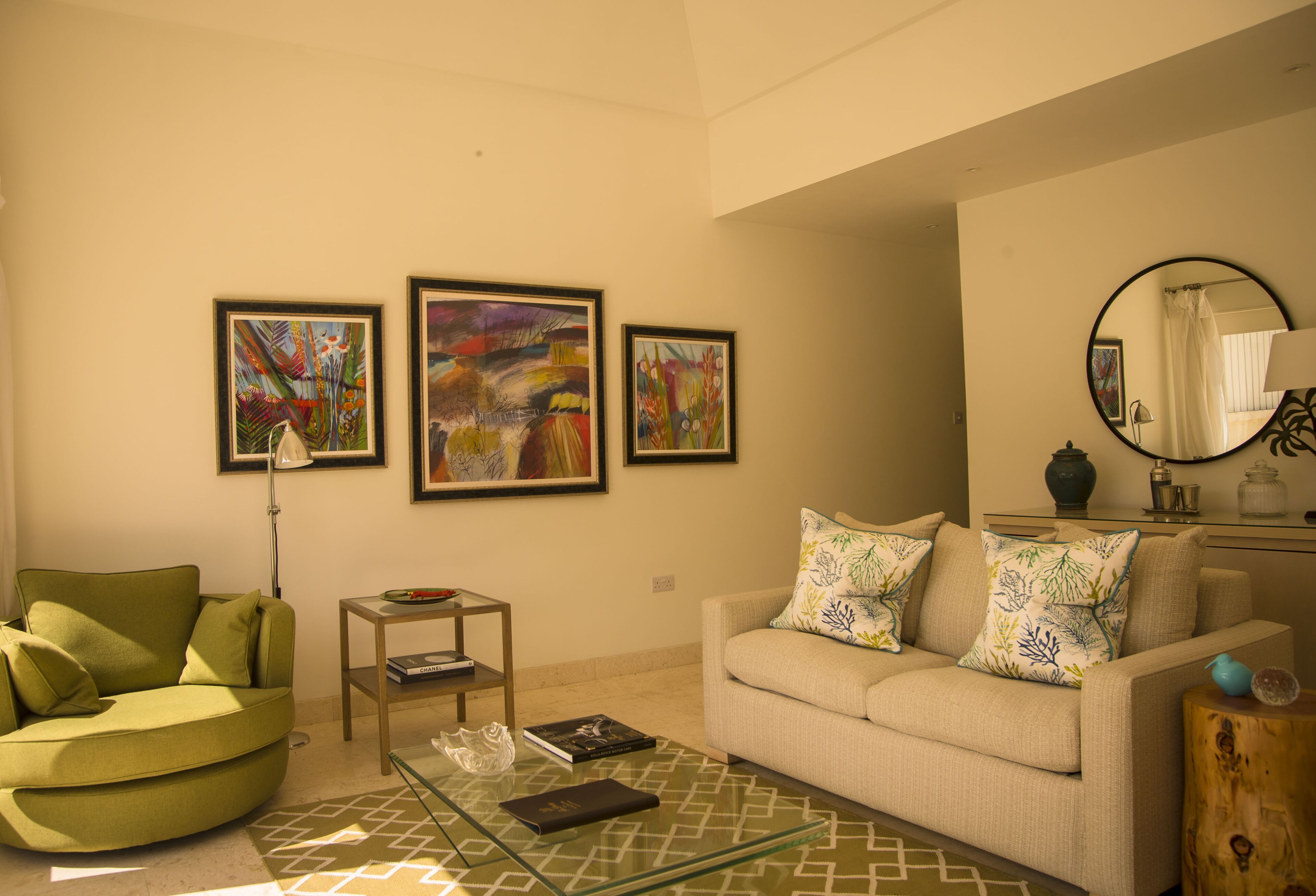 A living room with a green sofa, a beige sofa, and a glass coffee table. There are three framed artworks on the wall.