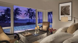 Un salon avec vue sur une plage au coucher du soleil, une table basse avec des bougies et un tableau encadré au mur.