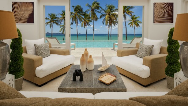 Un salon avec vue sur une plage et des palmiers. On y trouve deux canapés, une table basse et une maquette de voilier.