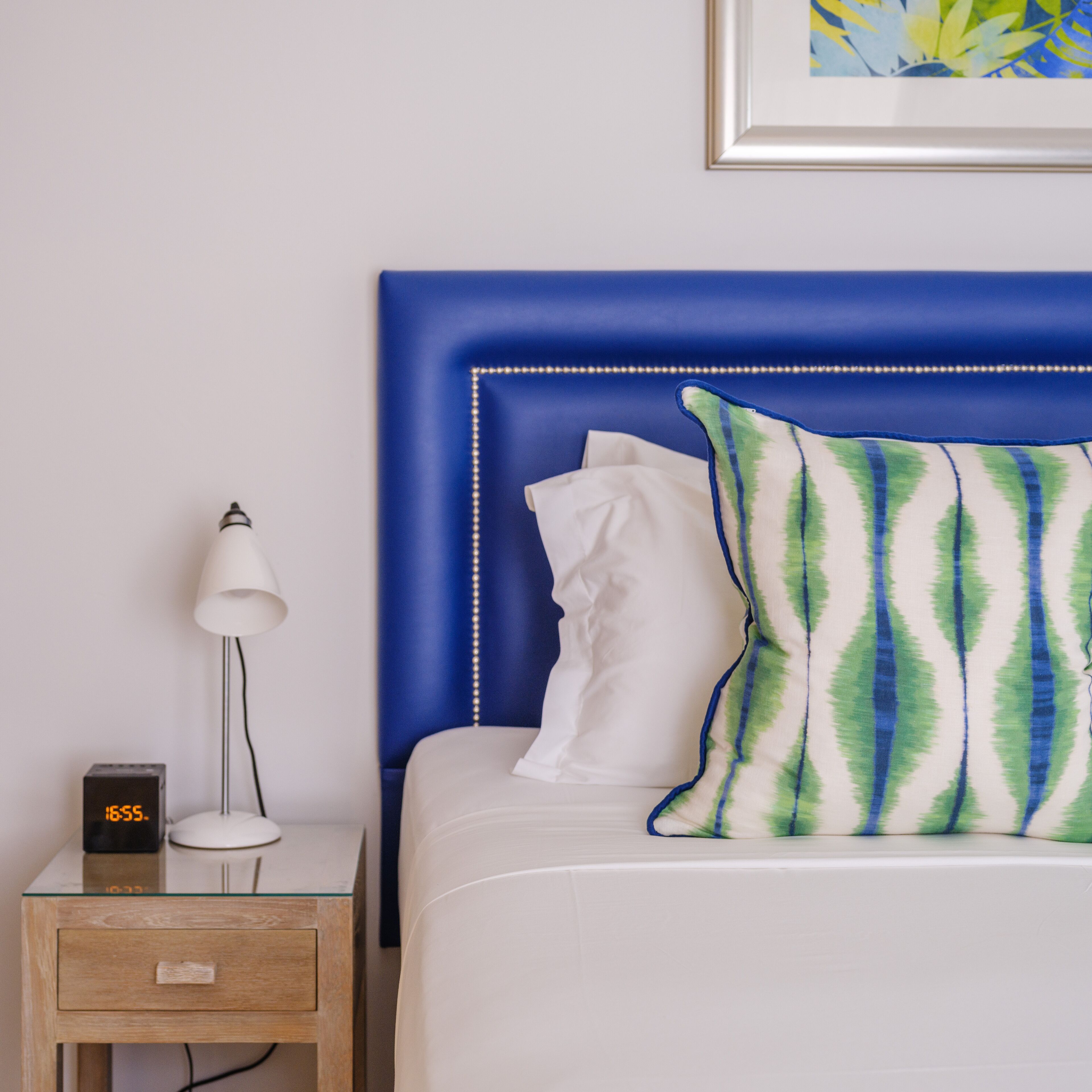 Una cama bien tendida con un cabecero azul, una almohada blanca y una almohada con estampado blanco y verde. Hay una mesita de noche con una lámpara y un reloj digital que muestra la hora.