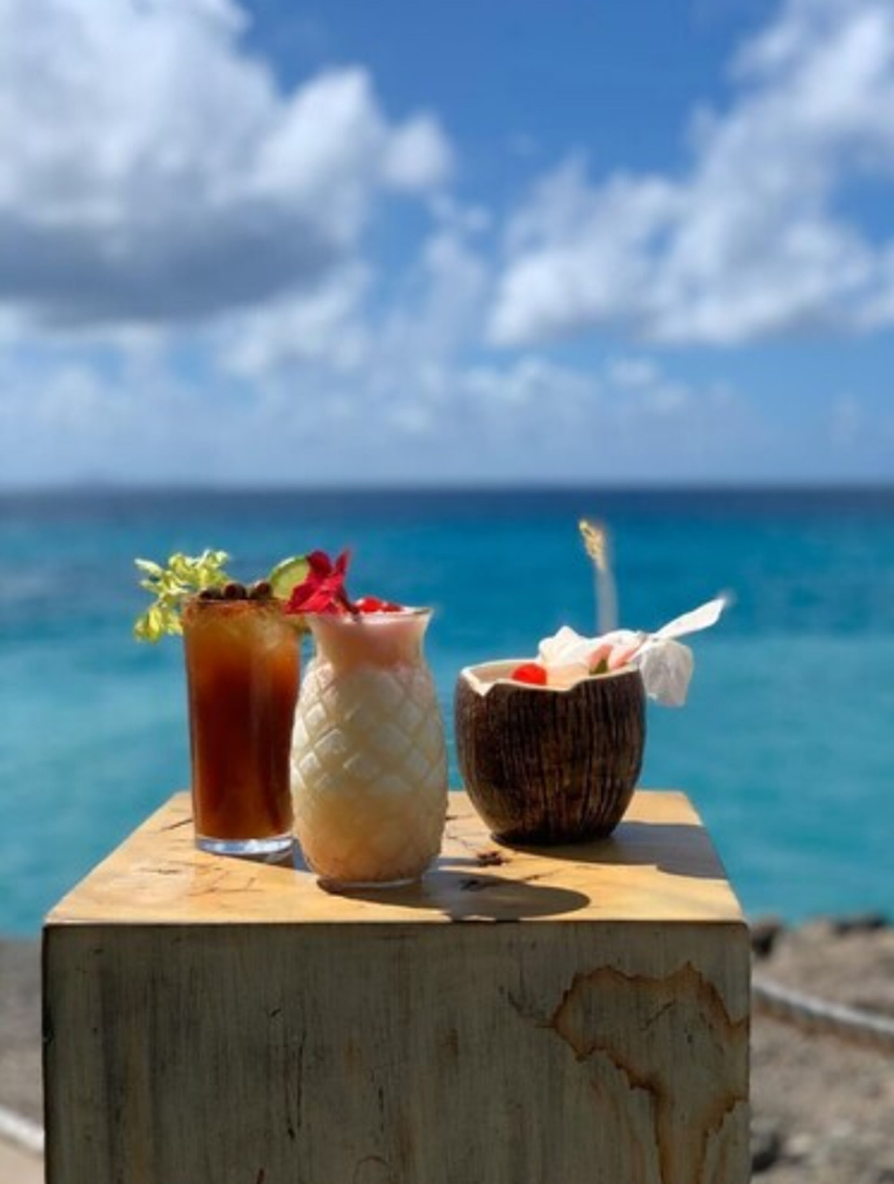 A wooden table with three drinks: a tall glass with a straw and garnish, a glass with a textured pattern, and a bowl with a straw and a wrapped item. The background features a clear blue sky and a body of water.