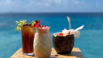 A wooden table with three drinks: a tall glass with a straw and garnish, a glass with a textured pattern, and a bowl with a straw and a wrapped item. The background features a clear blue sky and a body of water.