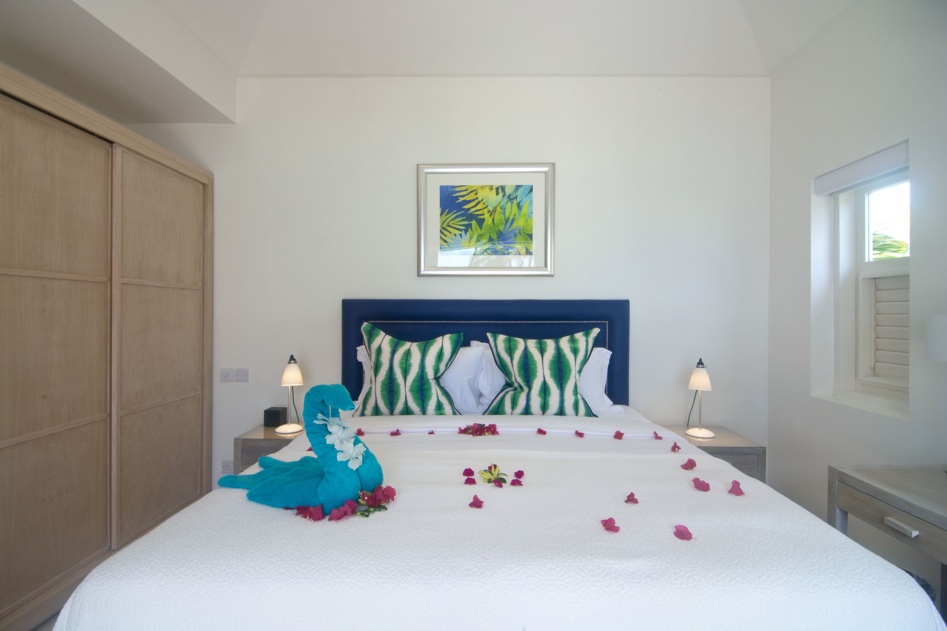 A neatly made bed with a blue blanket and pillows, a framed tropical artwork above it, and a window with shutters on the right.