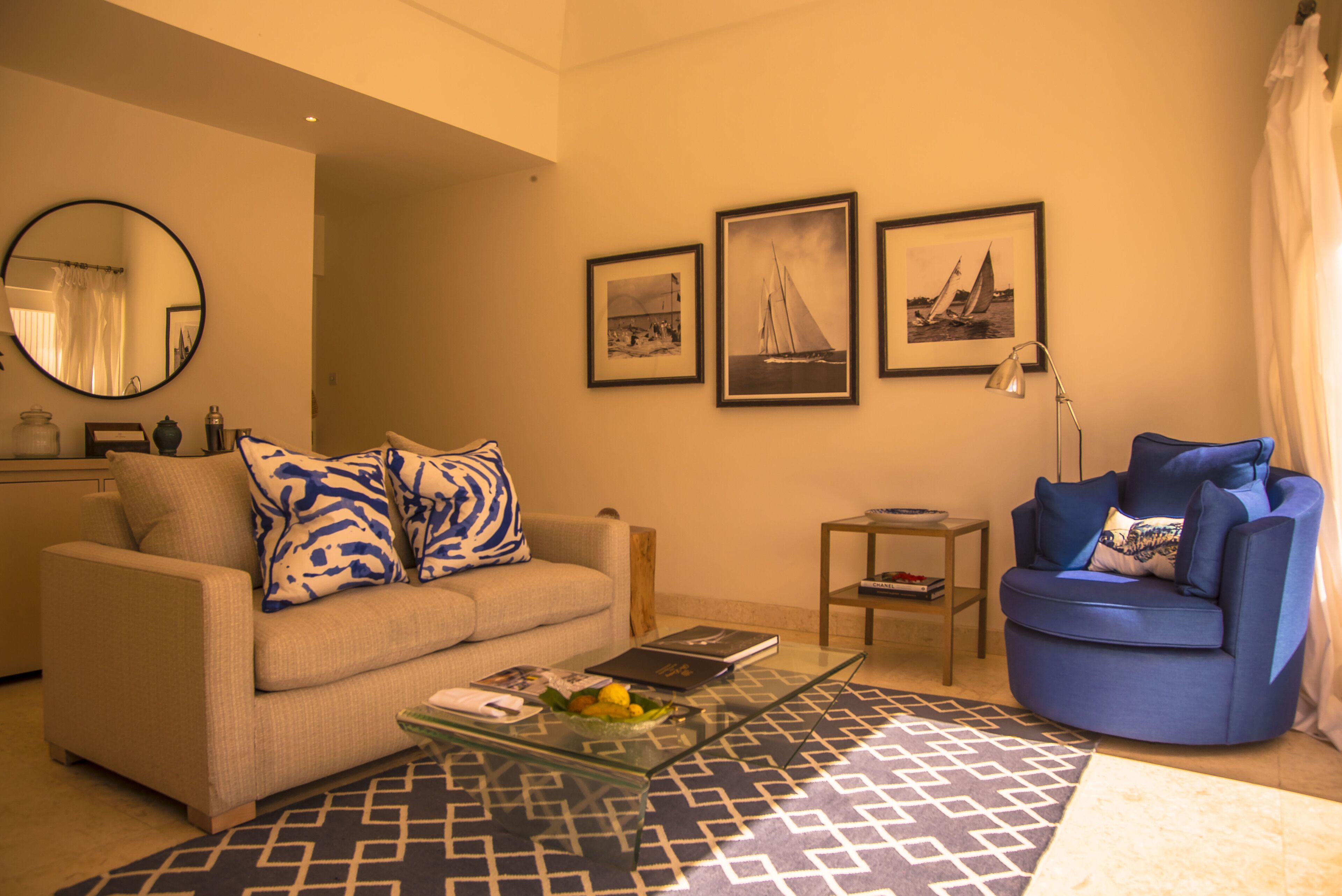 Un salon avec un canapé beige, un fauteuil bleu, une table basse en verre et un tapis à motifs. Trois tableaux encadrés ornent le mur, ainsi qu’un miroir rond et une petite table d’appoint avec une lampe.