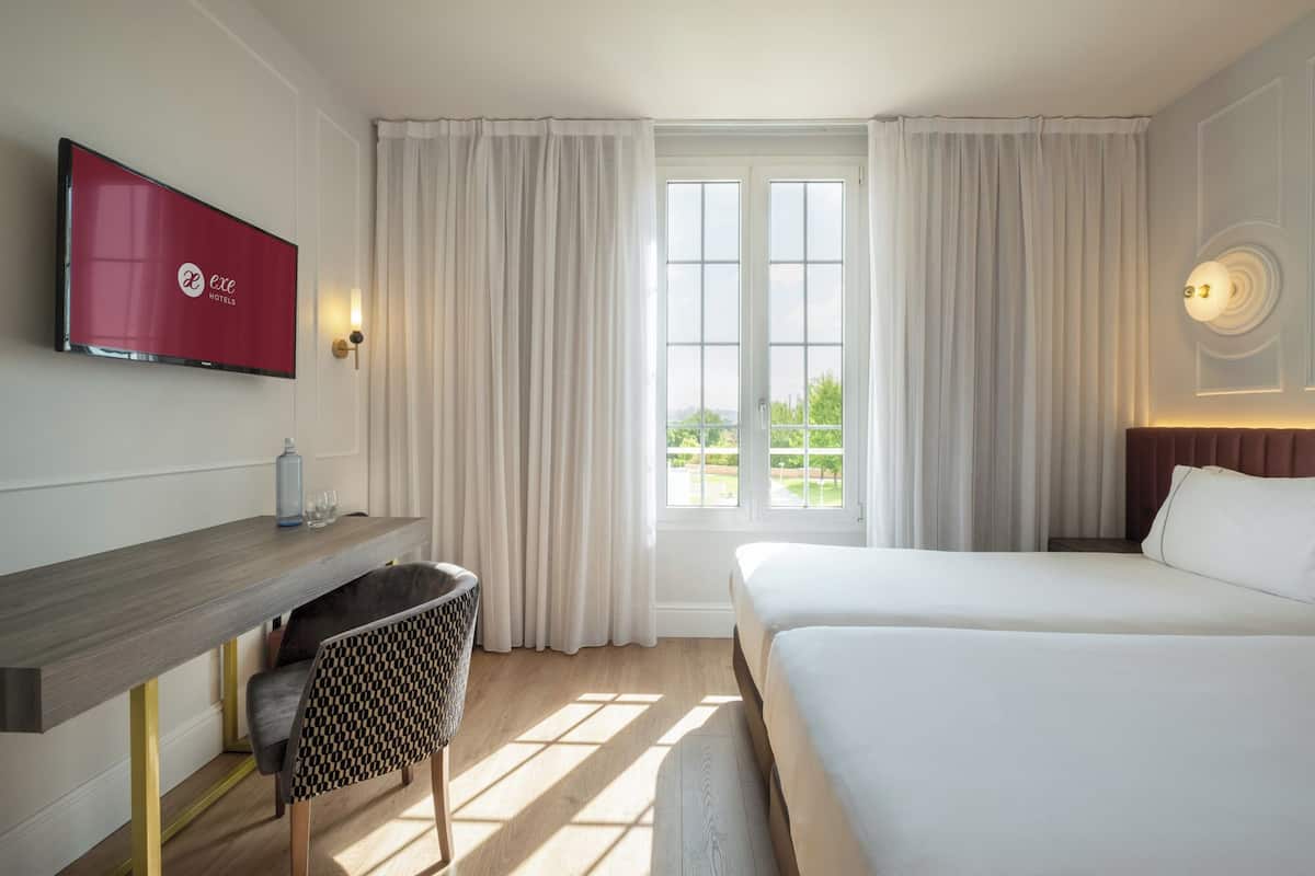 Une chambre d’hôtel avec un lit, un bureau, une chaise, une télévision et une fenêtre avec des rideaux.